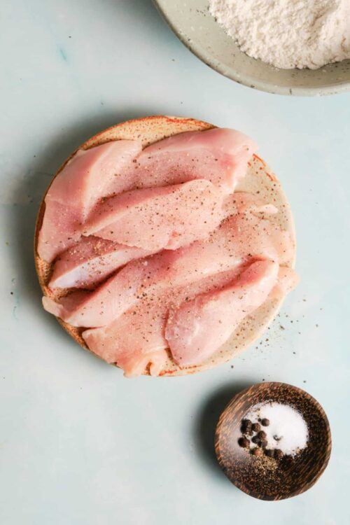 Sliced raw chicken breast seasoned with pepper and salt on a cutting board.