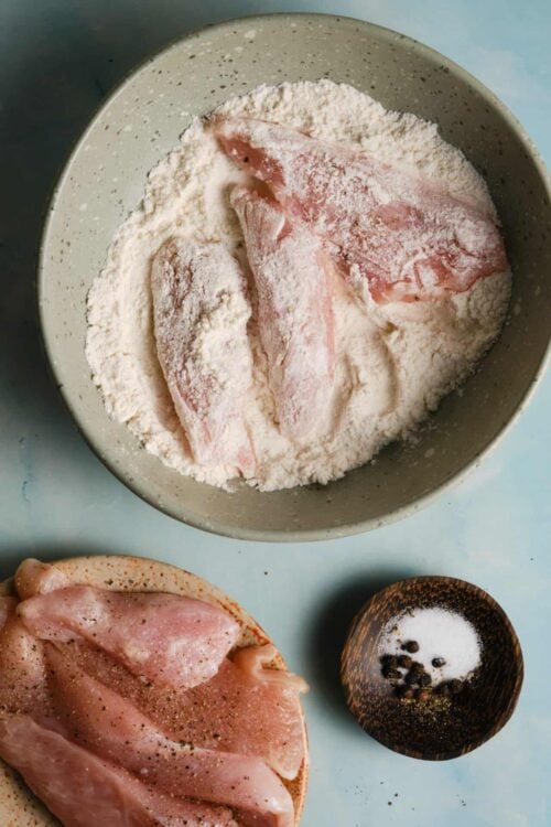 Flour-dusted raw chicken breasts in a mixing bowl with seasonings and peppercorns nearby.