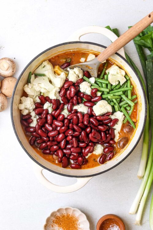 Fresh vegetable chili being prepared with red beans, cauliflower, and green beans in a white pot.