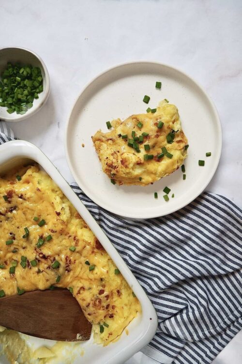 Creamy cheesy baked breakfast casserole with green onions in a white baking dish and on a white plate.