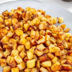 Crispy pan-fried potatoes served on a white plate with a bowl of red dipping sauce in the background.