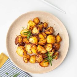 Golden roasted potatoes served on a beige plate with rosemary garnish, next to a fork and salt, perfect for a savory side dish.