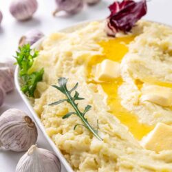 Creamy garlic mashed potatoes with butter and herbs on white platter.