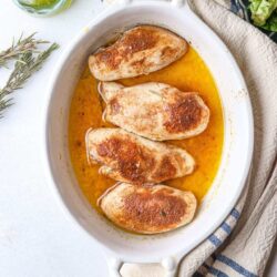 Juicy baked chicken breasts in a creamy sauce with herbs in a white baking dish.