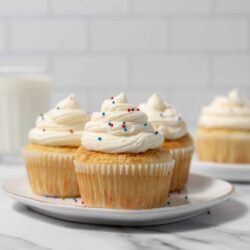 Buttery vanilla cupcakes with swirls of creamy frosting and colorful sprinkles on a white plate.