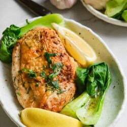 Juicy baked chicken breast with garlic, lemon, and fresh greens on a rustic white plate.