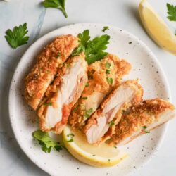 Crispy chicken parmesan cut into slices with lemon wedges and fresh parsley on a white plate.