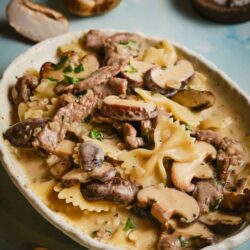 Creamy mushroom and beef stroganoff in a white dish on blue background with sliced mushrooms.