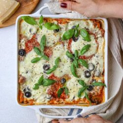 Savory baked lasagna with cheese, tomatoes, olives, and fresh basil in a white casserole dish.
