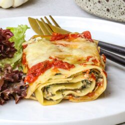 Roasted vegetable lasagna with cheese and tomato sauce on white plate.