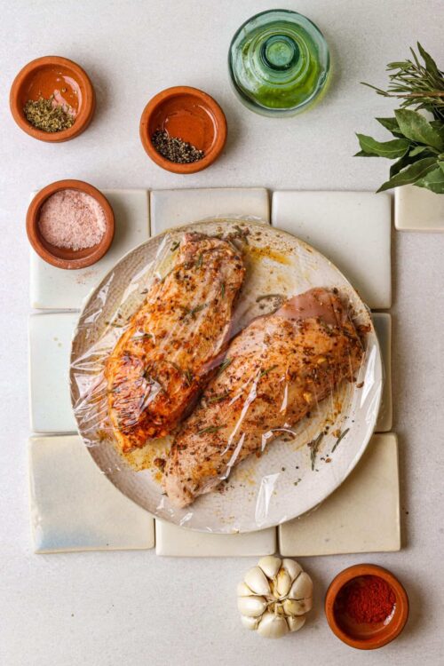 Juicy seasoned chicken breasts in plastic wrap with herbs and spices.