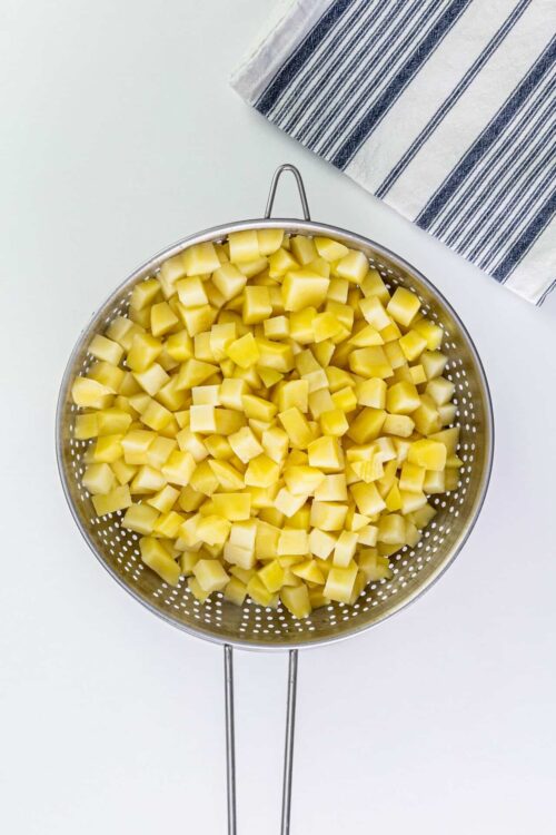Diced potatoes in a stainless steel colander over a white background.