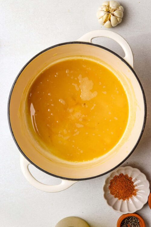 Creamy butternut squash soup in a white enameled pot with garlic, spices, and roasted vegetables around it.