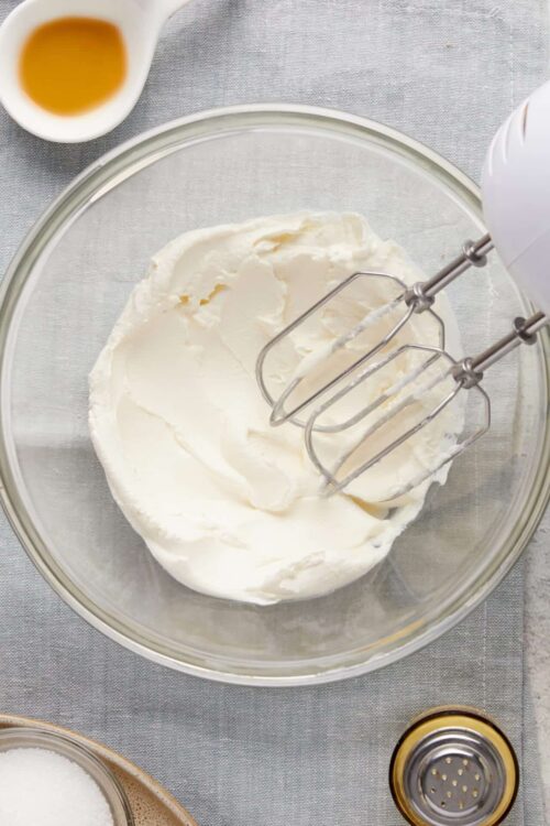 Fluffy whipped cream in a glass mixing bowl with hand mixer whisks. Baking ingredients nearby, ready for dessert or pastry preparation.