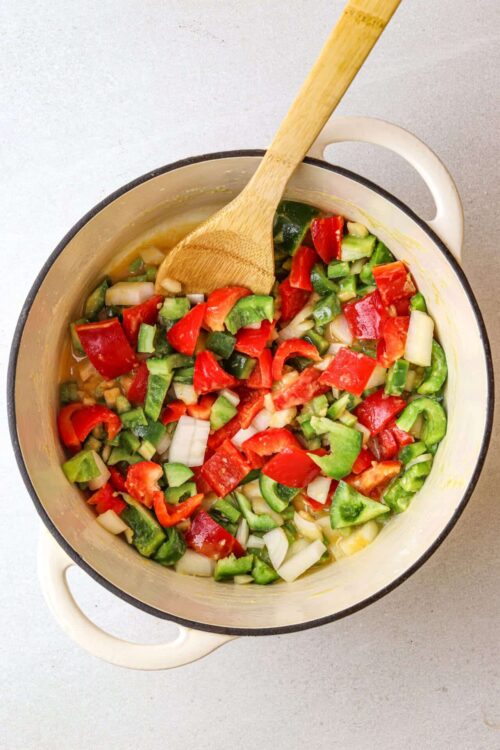 Diced colorful bell peppers and onions in a white enameled cast-iron pot with a wooden spoon.