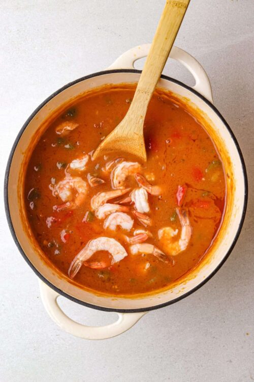 Savory shrimp and chili soup in a white enamel pot with a wooden spoon.