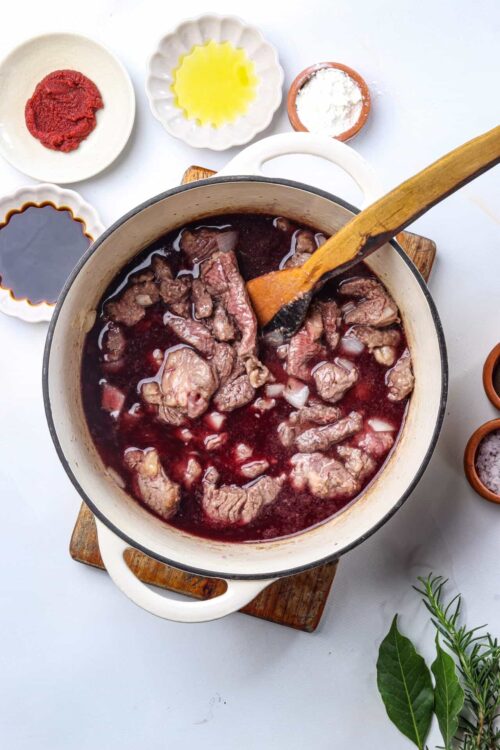 Braised beef stew cooking in a white Dutch oven with herbs and seasonings around it.
