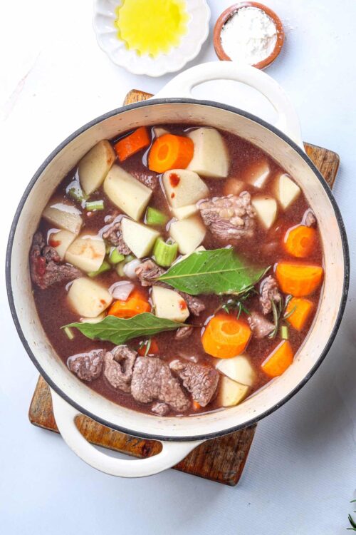 Hearty beef vegetable soup in a white enamel pot with carrots, potatoes, celery, bay leaves, and chunks of beef.