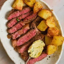 Juicy sliced steak with roasted potatoes and herb butter on a white plate.