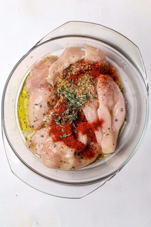 Boneless chicken breasts with herbs and spices in a glass baking dish.