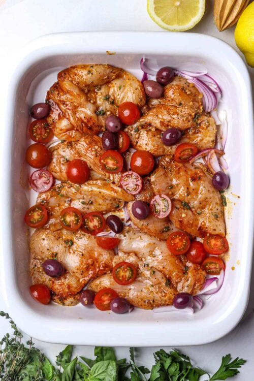 Juicy baked chicken thighs with cherry tomatoes, olives, and herbs in a white baking dish.