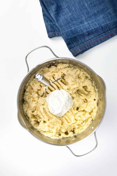 Butter and potato mashed in stainless steel mixing bowl with hand mixer, on white surface.