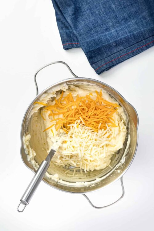 Shredded cheddar and Monterey Jack cheese layered on mashed potatoes in a stainless steel baking dish.
