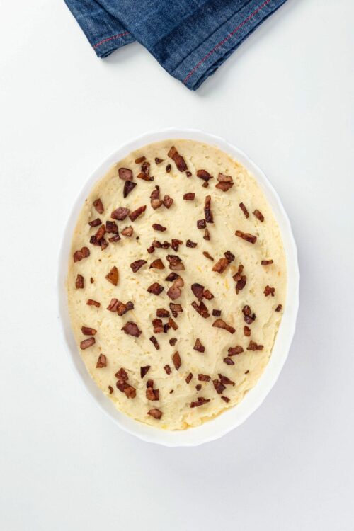 Creamy mashed potatoes with crispy bacon bits in a white bowl on a white surface.