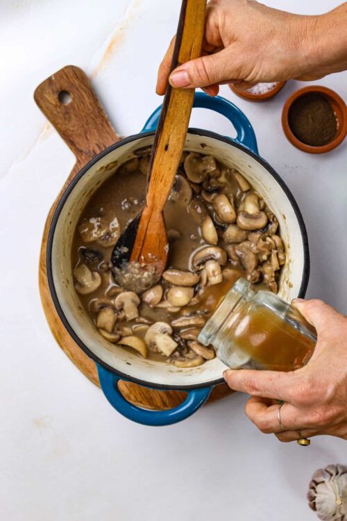 Creamy mushroom gravy simmering in a blue enamel pot with fresh mushrooms stirring in.