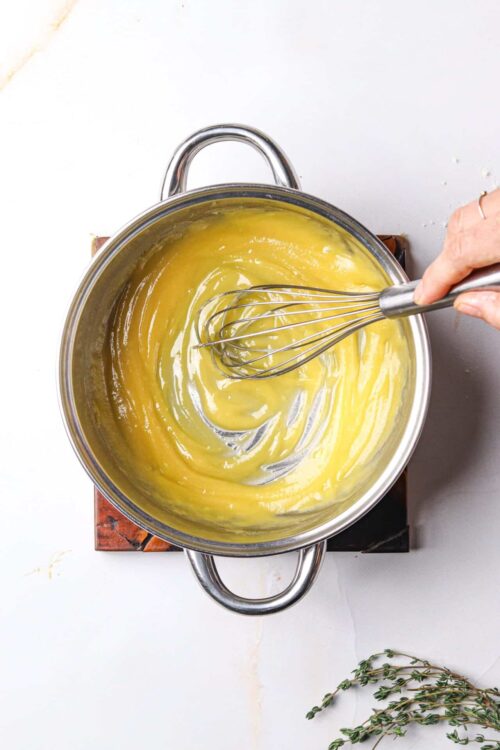 Silky lemon curd being whisked in a stainless steel pot for baking recipes.