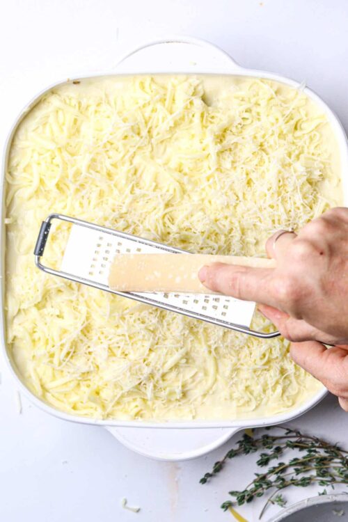 Shredded cheese being spread over a casserole dish with a grater, preparing a cheesy baked dish.