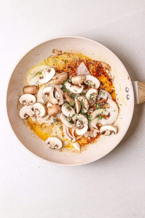 Sautéed mushrooms and onions cooking in a beige skillet with fresh herbs for a flavorful dish.