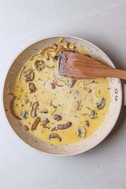 Sauteed mushroom and onion cream sauce in a beige skillet with a wooden spatula.
