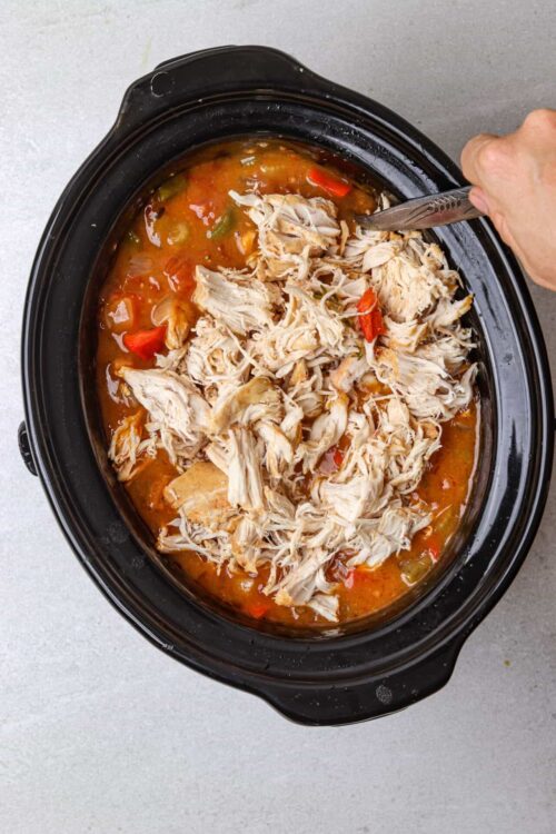 Shredded chicken being added to slow cooker with chicken and vegetable soup.