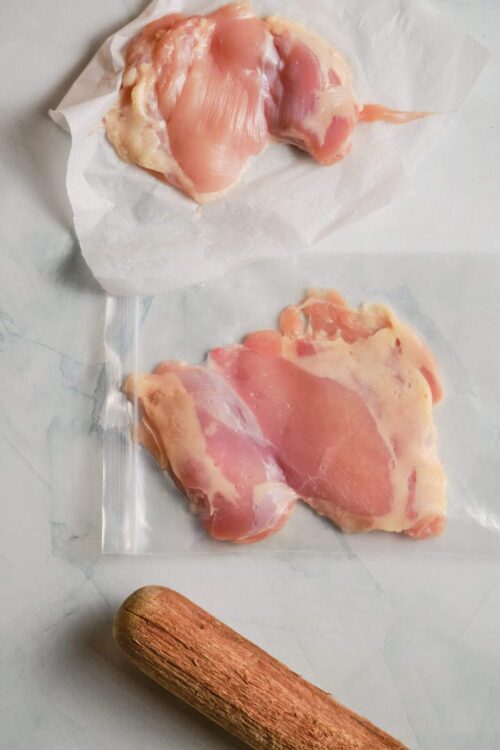 Raw chicken breasts on parchment paper with a rolling pin for cooking or baking preparation.