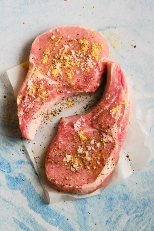Seasoned raw pork chops with salt, pepper, and herbs on parchment paper.