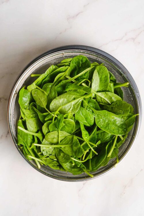 Fresh spinach leaves in a stainless steel strainer on white marble surface.