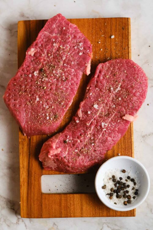 Juicy raw steaks seasoned with salt and pepper on a wooden cutting board.