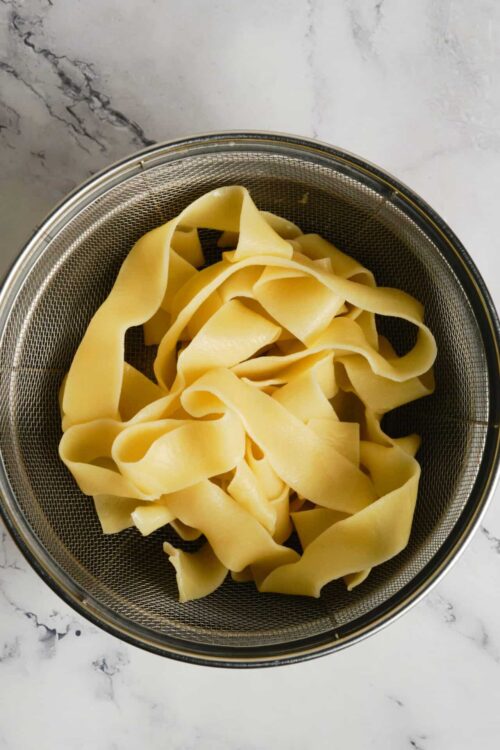 Fettuccine pasta in a metal strainer on marble surface.