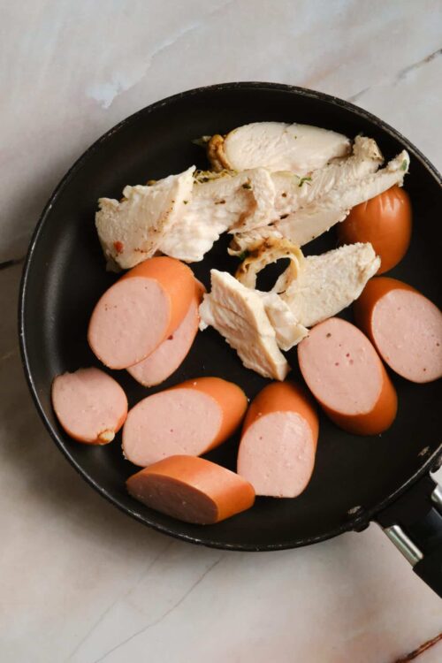 Sliced sausages and cooked chicken breast in a black skillet on a marble countertop.