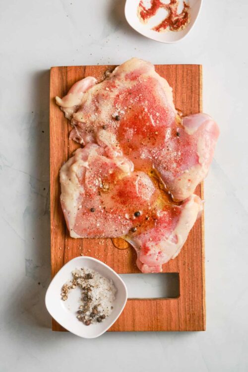 Raw chicken thighs seasoned with salt and pepper on a wooden cutting board.
