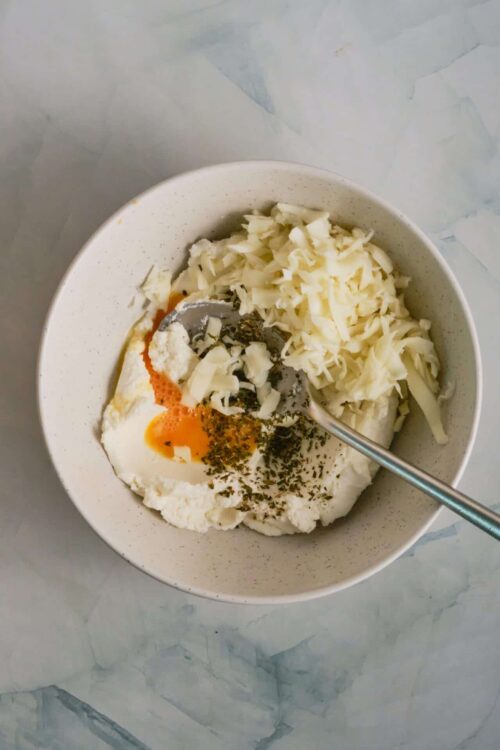 Cream cheese mixture for baking with herbs and shredded cheese in a white bowl.