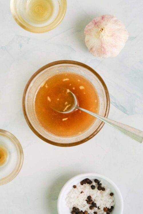 Golden garlic infusion in a glass jar with garlic bulb and salt pepper in a white bowl, on a marble surface.