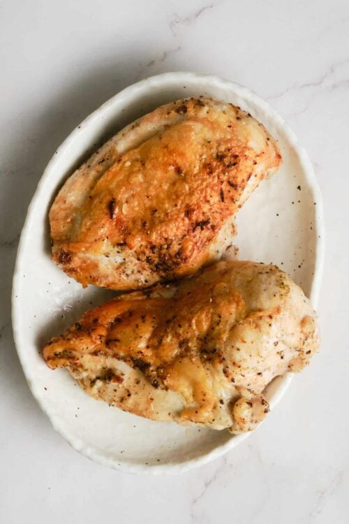 Juicy baked chicken breasts with golden crispy skin on a white ceramic dish, cooked to perfection.