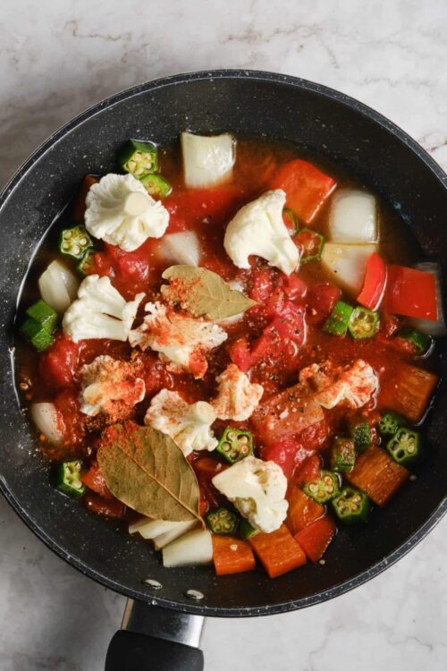 Fresh vegetable soup cooking in a black pot with tomatoes, cauliflower, okra, and herbs for healthy home-cooked meal.