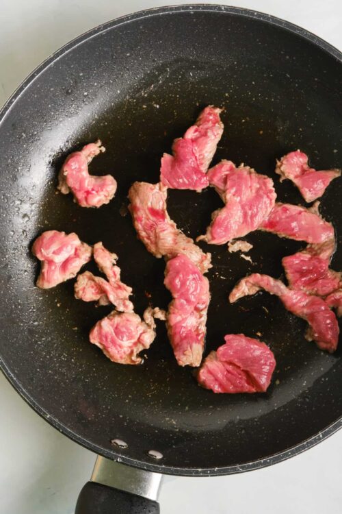 Sliced raw beef cooking in a black non-stick skillet.