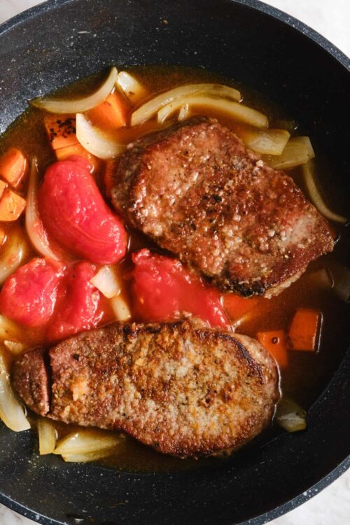 Juicy seared steak cooking with onions, carrots, and tomatoes in a skillet.