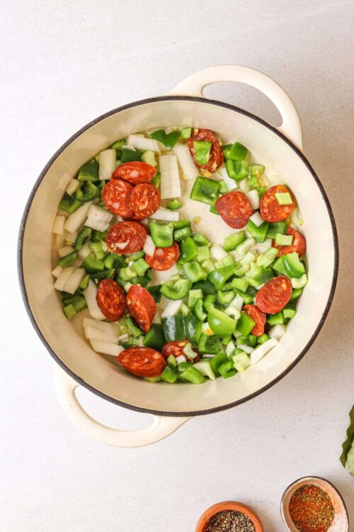 Diced green bell peppers, celery, onions, and pepperoni in a white cast iron skillet, ready for cooking.
