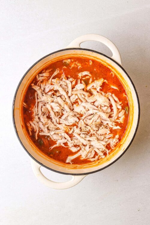 Shredded chicken in spicy tomato soup in a white enamel pot.