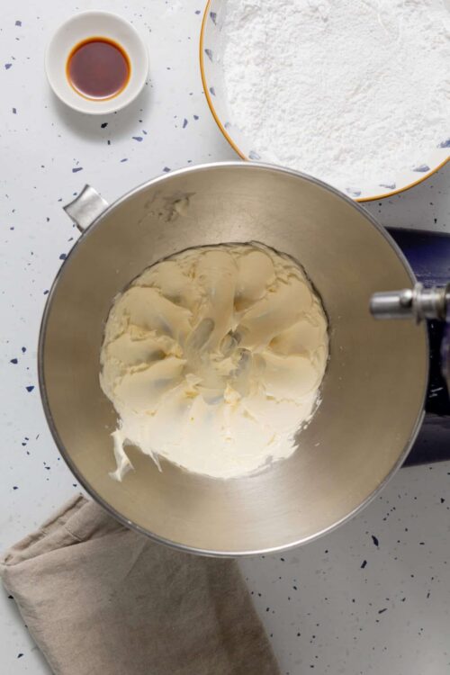 Cream cheese frosting in stand mixer bowl, ingredients for baking, baking supplies, baked goods, frosting preparation.